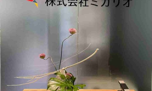 木製カウンターに飾られた、ピンクの丹頂アリウム、モンステラの葉、白いサラシの枝を使用したモダンなフラワーアレンジメント