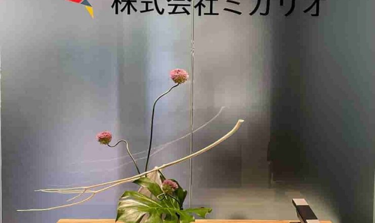 木製カウンターに飾られた、ピンクの丹頂アリウム、モンステラの葉、白いサラシの枝を使用したモダンなフラワーアレンジメント