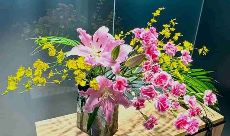 木製カウンターに置かれたガラス花瓶に、ピンクのスプレーカーネーション、ユリ、黄色いオンシジュウム、アレカヤシの葉を使用した華やかなフラワーアレンジメント