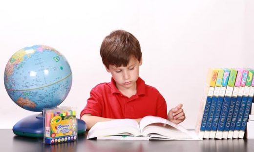 赤いポロシャツを着た少年が机に向かって本を読んでいる様子。机の上には青い台座の地球儀、クレヨンのセット、複数の参考書が並んでおり、白い背景で撮影されている