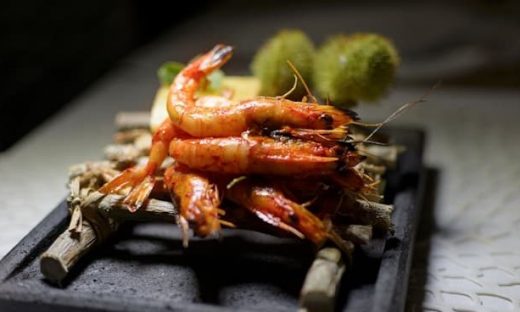 黒い長方形の皿に盛られた焼きエビ。竹の枝の上に数尾の大きな海老が重ねられ、鮮やかな赤色に焼き上がっている。添え物として緑色の野菜が添えられている
